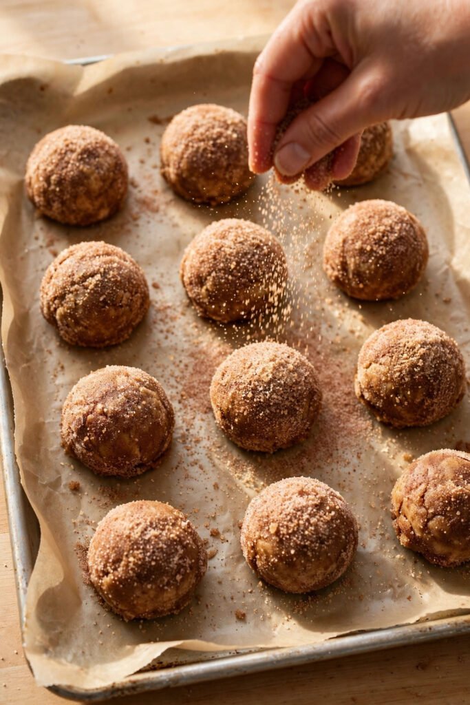 Snickerdoodle cookie dough balls being rolled in cinnamon sugar on a baking sheet